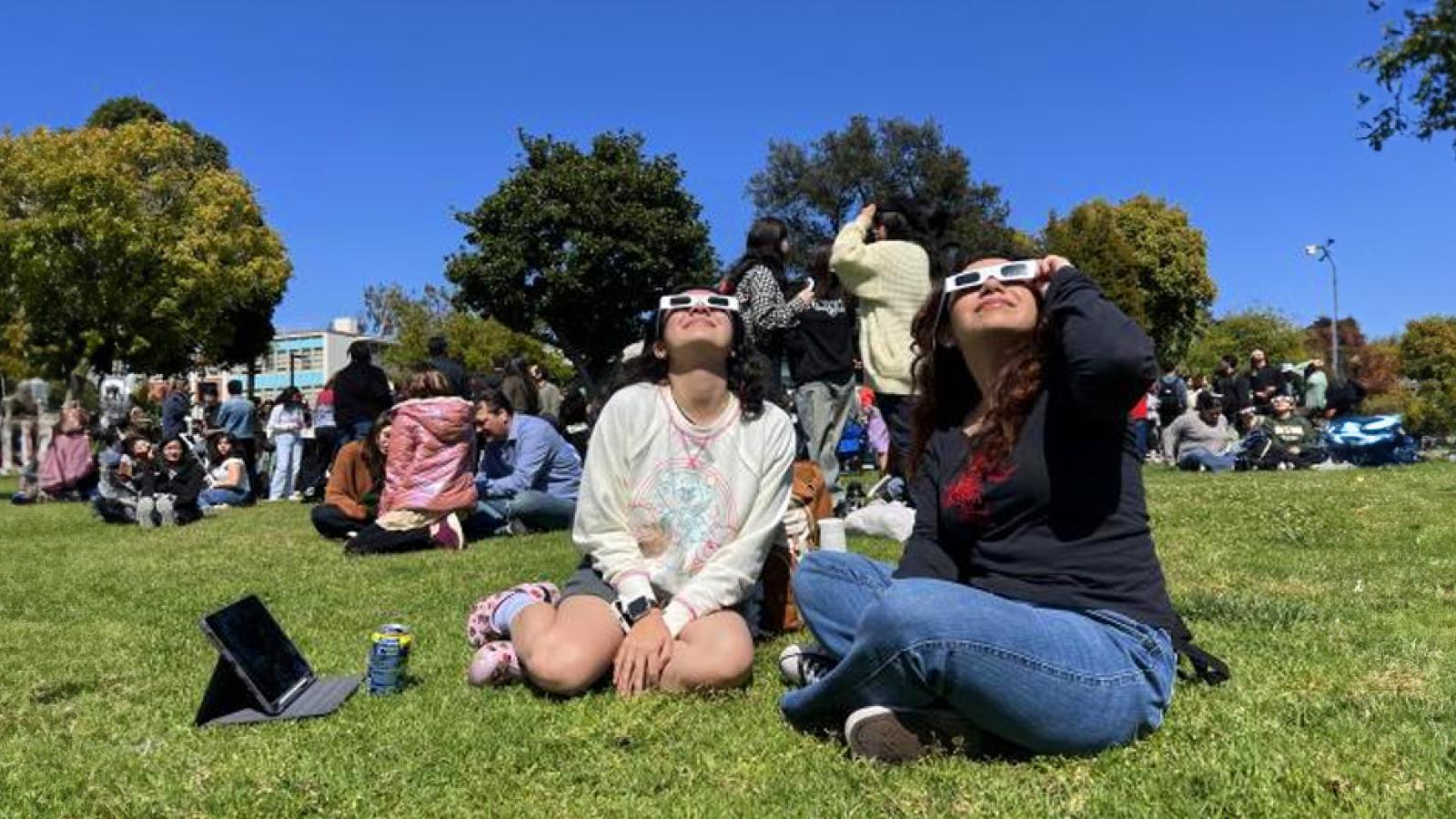 people sitting on grass watching eclipse with viewing glasses